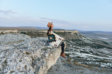 Wall Mural - Serene woman in hat and blanket sitting on rock, gazing at stunning mountain valley view