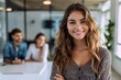 © Pinklife - A joyful young woman with wavy hair smiles brightly, with colleagues engaged in conversation in a blurry background