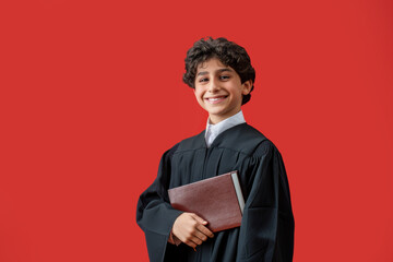 Smiling boy in graduation robe holding a book against red background