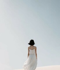 Wall Mural - A woman wearing a white dress is standing with her back turned to the camera. The background is a clear blue sky.

