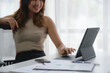 © amnaj - Smiling woman working on a tablet at a modern workspace. The desk has documents and a phone, with natural light filtering through the window.