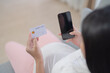 © tampatra - A portrait of happy Asian pregnant woman using a mobile phone device with credit card, shopping online in bedroom at home. Having a baby. Family people lifestyle. Mom love.