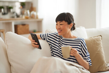 An Asian woman using mobile phone while enjoying coffee at home