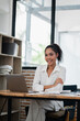 © Satori Studio - Young professional woman smiling at desk in modern office with laptop and bookshelves in background.