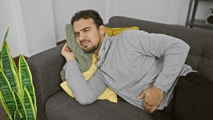 Wall Mural - Young hispanic man with beard feeling unwell, clutching stomach, lying on couch indoors.