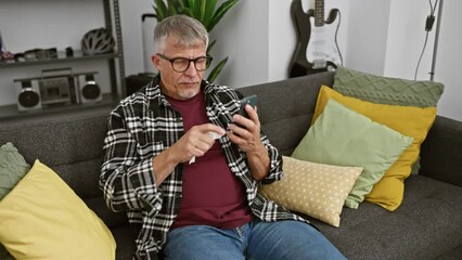 Poster - Grey-haired man texting and talking on a smartphone in a modern living room, showing casual home life.