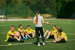 © master1305 - Youth Sports Education. Group of enthusiastic young soccer players listening to strategic advice from female coach, focusing on teamwork and skill development. Sport, school, active lifestyle concept
