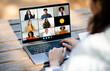 © Prostock-studio - Unrecognizable woman sitting at a desk, typing on a laptop while video conferencing with a diverse group of people displayed on the screen. The individuals on screen are engaged in a virtual meeting