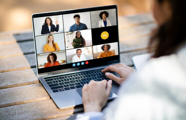 Poster - Unrecognizable woman sitting at a desk, typing on a laptop while video conferencing with a diverse group of people displayed on the screen. The individuals on screen are engaged in a virtual meeting