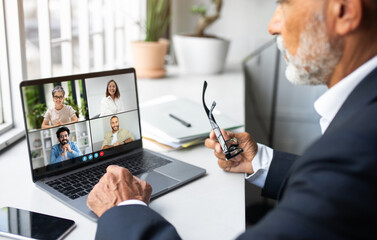 Poster - A mature businessman is engaged in a virtual meeting with colleagues via his laptop in a bright and modern office setting. He is holding his glasses