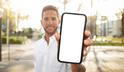 Fototapete - A man in a white shirt stands outdoors, holding a smartphone in front of him with a blank white screen. He is smiling and looking directly at the camera.