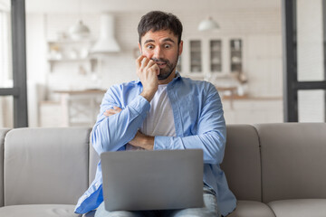 Poster - Man sitting on a couch in a home setting. He is looking at his laptop with a worried expression on his face and has his hands clasped together in front of him. He appears to be stressed or anxious.
