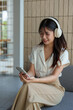 © Phimwilai - Happy Asian woman listening to music on mobile phone while sitting on sofa at coffee shop Relaxed smiling young woman with headphones in the morning Holiday relaxation concept.