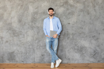 Poster - Man standing in front of a concrete wall, leaning against it with one hand in his pocket. He is smiling at the camera and wearing headphones over his ears. He is holding a laptop in his other hand