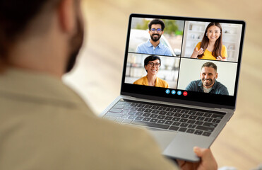 Poster - A man sitting at a table is using a laptop, which displays a group of people on the screen. He appears engaged in a virtual meeting or video conference with his colleagues.