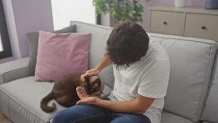 Wall Mural - A young man caresses a brown siamese cat while sitting on a gray sofa in a cozy living room.