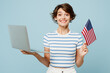 © ViDi Studio - Young surprised IT woman wears striped t-shirt casual clothes hold American flag use work on laptop pc computer isolated on plain pastel light blue cyan background studio portrait. Lifestyle concept.