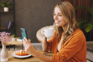 Wall Mural - Young smiling happy woman wear orange shirt do selfie shot on mobile cell phone talk speak sits alone at table in coffee shop cafe restaurant indoors work or study. Freelance office business concept.