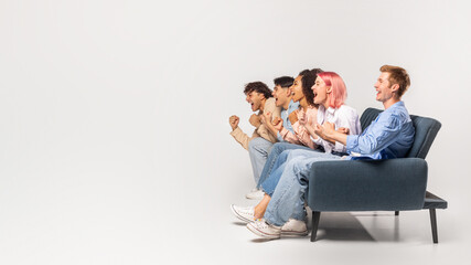 Five diverse young friends are sitting on a couch in a studio setting. They are all looking off to the side, cheering excitedly. They are likely watching a sporting event or a game show, copy space