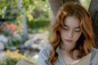 © Irina B - Young red-haired girl reading a book outdoors under a tree with a peaceful garden in the background..