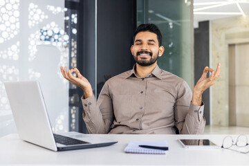 Relaxed Indian young man sitting in lotus position in chair at desk in office and meditating. He rests after closing his eyes.