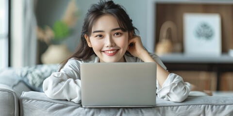 Wall Mural - Smiling woman with dark hair working on a laptop, lying on a couch, in a bright, cozy room.