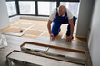 © anatoliy_gleb - Man in work overalls installing laminate timber flooring in room with window. Male construction worker laying laminate wood plank on floor underlayment. Hardwood floor renovation concept.