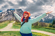 © EdNurg - A woman in a jacket and orange hat is standing in a field with mountains in the background. She is smiling and enjoying the beautiful scenery