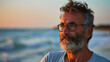 © Nataliia_Trushchenko - A man with a beard and glasses is standing on a beach, looking out at the ocean. The sky is a beautiful mix of orange and blue, creating a serene and peaceful atmosphere. The man is enjoying the view