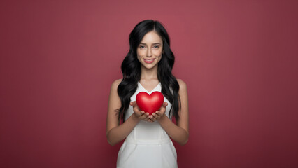 Smiling woman holding a red heart-shaped object, standing against a red background, symbolizing love, care, and affection.