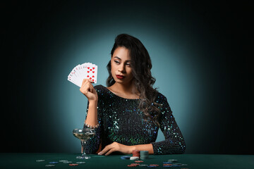 Wall Mural - Beautiful young African-American woman sitting at poker table with playing cards, chips and glass of champagne on dark green background