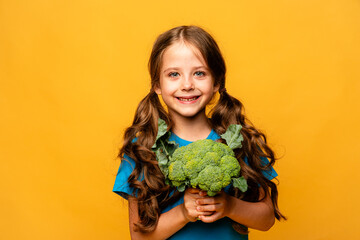 Happy smiling little girl in blue t-shirt standing with green raw broccoli over yellow background. Healthy food for children concept