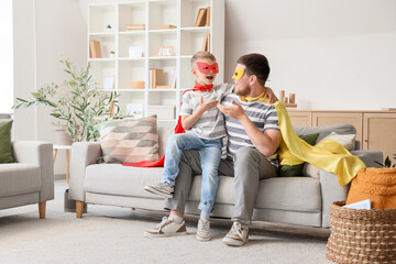 Wall Mural - Little boy with his father in superhero costumes talking on sofa at home