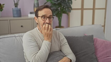 Wall Mural - Middle-aged man with glasses looks surprised while sitting on a grey couch inside a cozy living room, holding a remote.