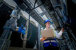 © ultramansk - An industrial worker in high-visibility gear and a blue helmet meticulously examines the intricate network of machinery. The engineer is dedicated to maintaining operational efficiency and safety.