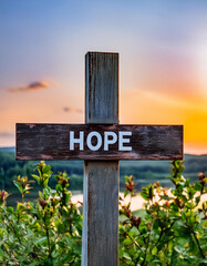 Wall Mural - A wooden cross sign with the word HOPE written on it, with a blurred background of nature and sky at sunset