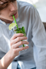 Wall Mural - Teenage boy in light blue shirt drinking refreshing summer iced cocktail, holding glass with mojito drink in hand, lifestyle, soft natural sunlight