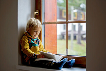 Wall Mural - Cute toddler child, blond boy, sitting on window, reading