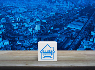 Wall Mural - Hammer and wrench with house icon on white block cube on wooden table over modern city tower, street, expressway and skyscraper, Business home service concept