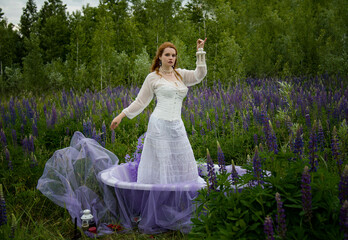 beautiful romantic readhead bride in vintage white wedding dress staying in bath the field of lupines flowers in the summer evening and holding lantern