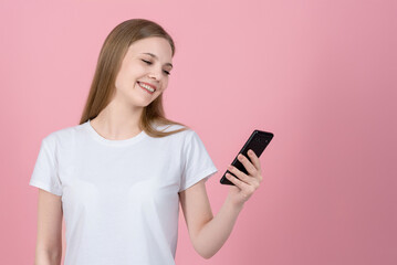 Wall Mural - Attractive caucasian young blonde woman in casual white t-shirt smiling and using mobile phone isolated on pink studio background. Wireless, app, internet, social media, online.