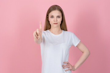 Wall Mural - Attractive serious caucasian young blonde woman in casual white t-shirt showing stop, one finger gesture, taboo sign isolated on pink studio background.