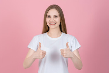 Wall Mural - Attractive caucasian young blonde woman in casual white t-shirt smiling, looking at camera and showing thumbs up isolated on pink studio background. Agreement, Good, Success.