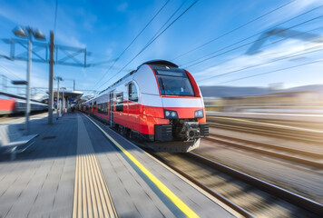 High-speed red passenger train moving at railway station platform under clear blue sky at sunset. Train station. Modern railway transportation concept with blurred motion effect. Railroad. Commercial