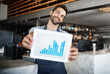 © peopleimages.com - Happy man, portrait and tablet screen with chart for profit, revenue or statistics at cafe. Young male person, waiter or business owner with smile for graph, growth or technology at indoor restaurant
