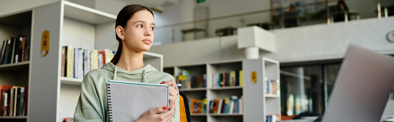 Wall Mural - A teenage girl stands in front of a bookcase in a library, immersed in studying and doing homework on her laptop after school.