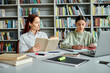 © LIGHTFIELD STUDIOS - a redhead tutor and a teenage girl, sit at a library table. The tutor teaches while using a laptop for modern education.