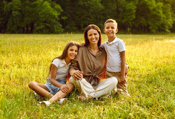 Wall Mural - Happy woman with daughter and son sitting in the field on green grass and looking cheerful at camera. Smiling mother hugging her two children in the summer park. Family leisure concept.