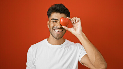 Wall Mural - Handsome young hispanic man with beard smiling and holding a heart over red isolated background