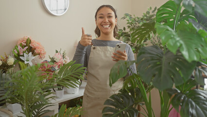 Sticker - A cheerful young hispanic woman gives a thumbs-up in a flower shop with a smartphone in hand.